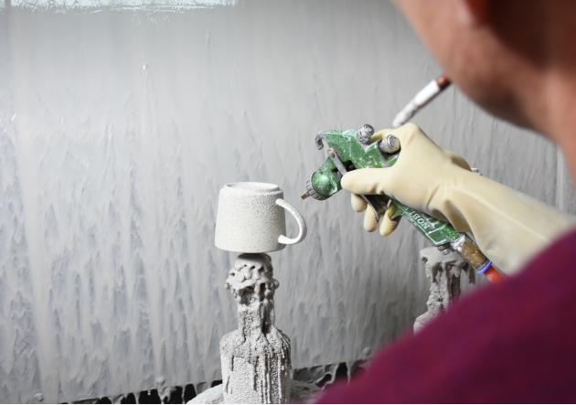 A worker holds a spray gun to spray glaze on a mug..png